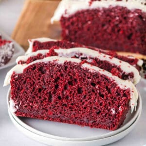 Square image of sliced red velvet loaf with cream cheese frosting on a plate, showing the red interior and white frosting.