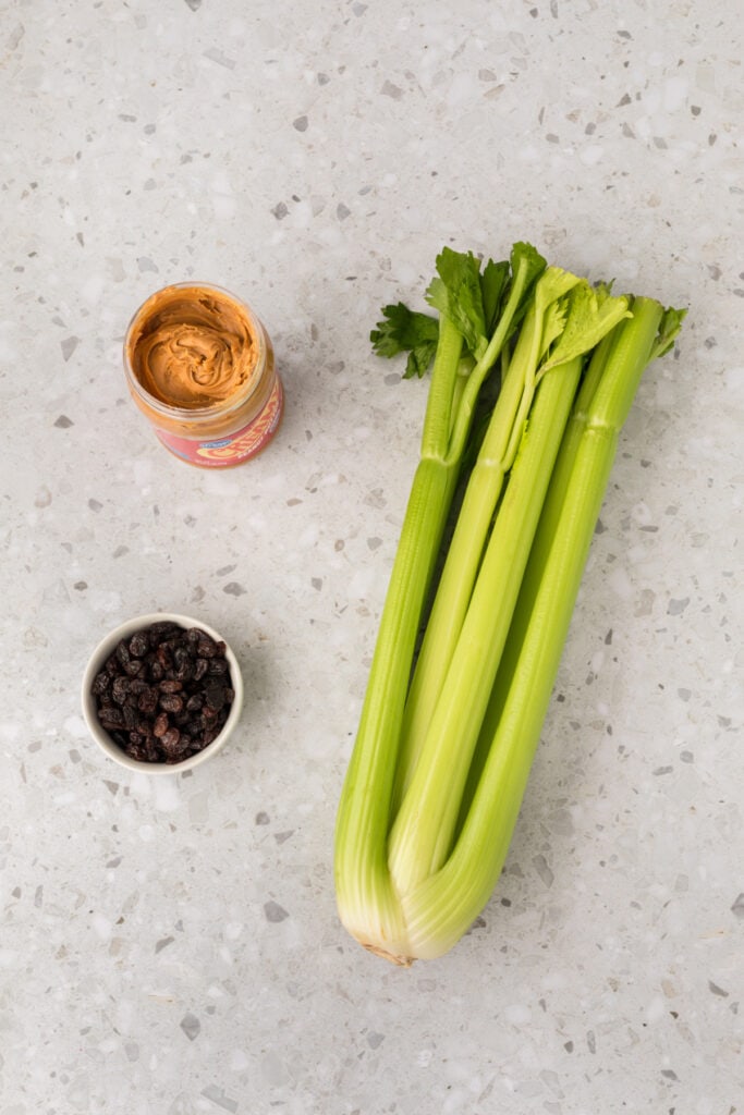 whole celery stalks on a countertop with a jar of peanut butter and a small bowl of raisins