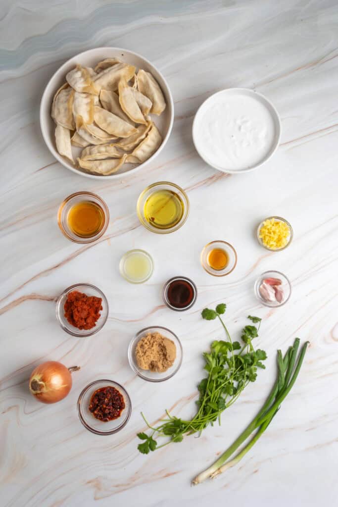 overhead view of frozen dumplings, coconut milk, curry paste, garlic, onion, ginger, cilantro, green onions, chili crunch, and small bowls of sauces arranged on a marble surface