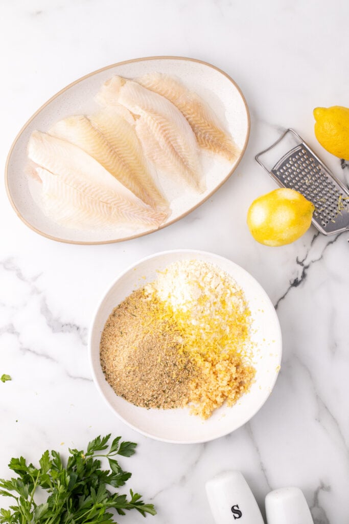 raw flounder fillets on a plate next to a bowl with breadcrumbs, parmesan, garlic, and lemon zest with lemons and a grater nearby