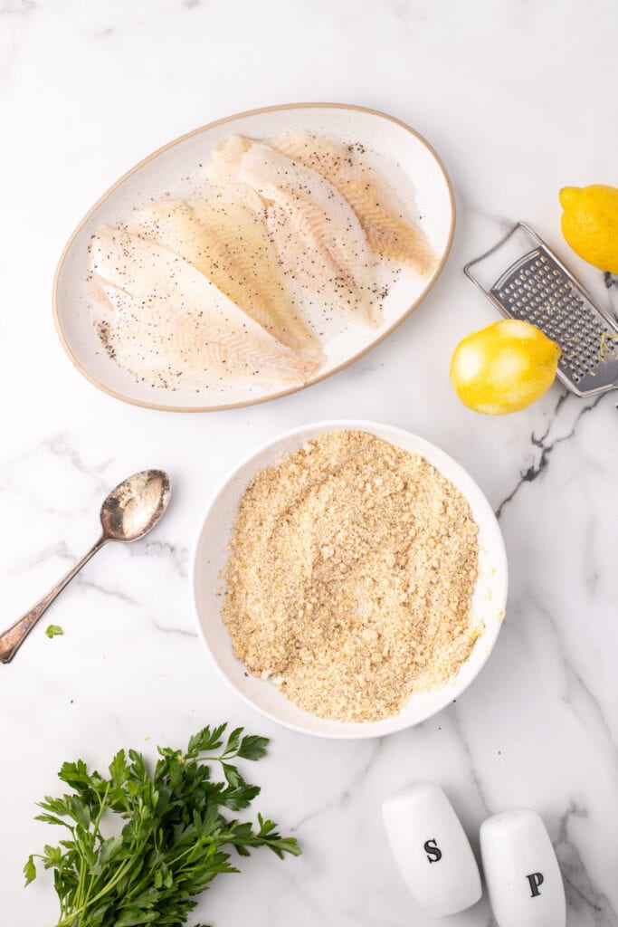 raw flounder fillets seasoned with black pepper next to a bowl of breadcrumb and parmesan mixture with parsley and lemons