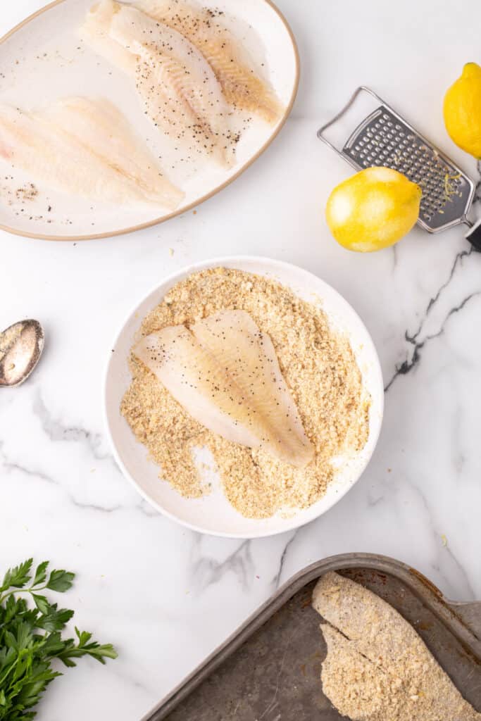flounder fillet being coated in breadcrumb and parmesan mixture in a bowl with lemons and grater nearby