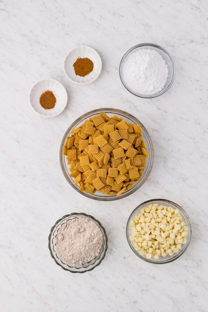 ingredients for carrot cake muddy buddies including Corn Chex cereal, white chocolate chips, powdered sugar, carrot cake mix, cinnamon, and nutmeg in bowls