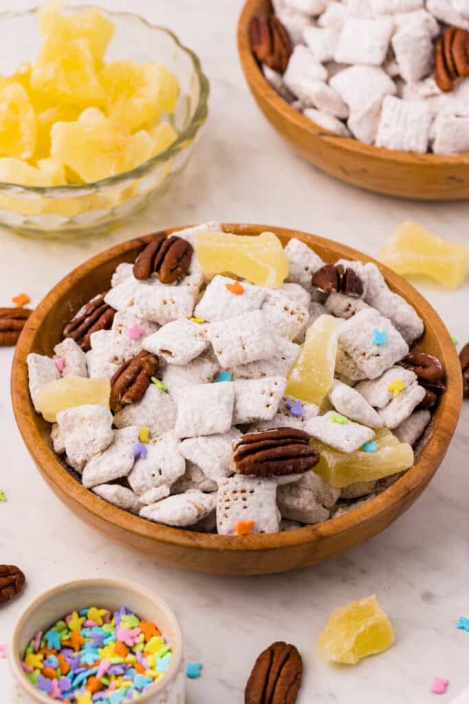 carrot cake muddy buddies in a wooden bowl with pecans, pineapple chunks, and pastel sprinkles