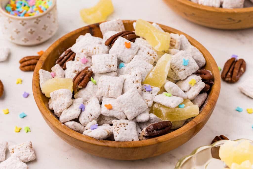 carrot cake muddy buddies in a wooden bowl with pecans, pineapple pieces, and colorful sprinkles on a countertop