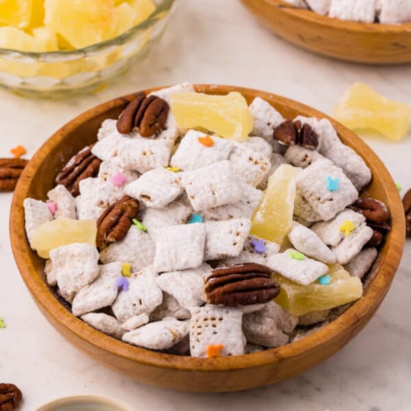 square image of carrot cake muddy buddies in a wooden bowl with pecans, pineapple chunks, and pastel sprinkles