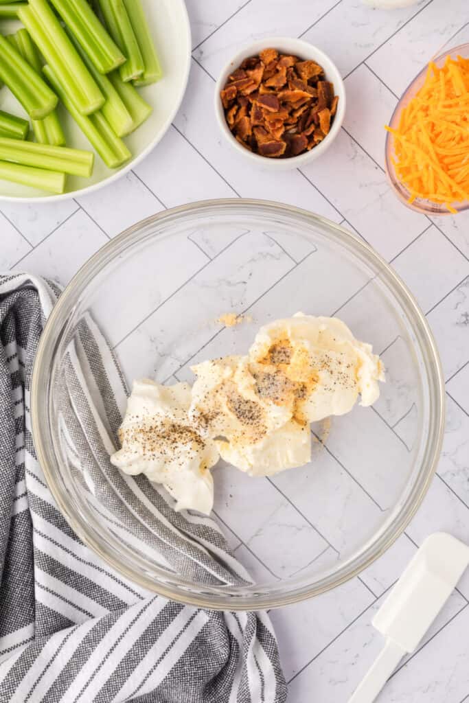 cream cheese and sour cream with seasonings in a glass mixing bowl next to celery, bacon, and shredded cheddar