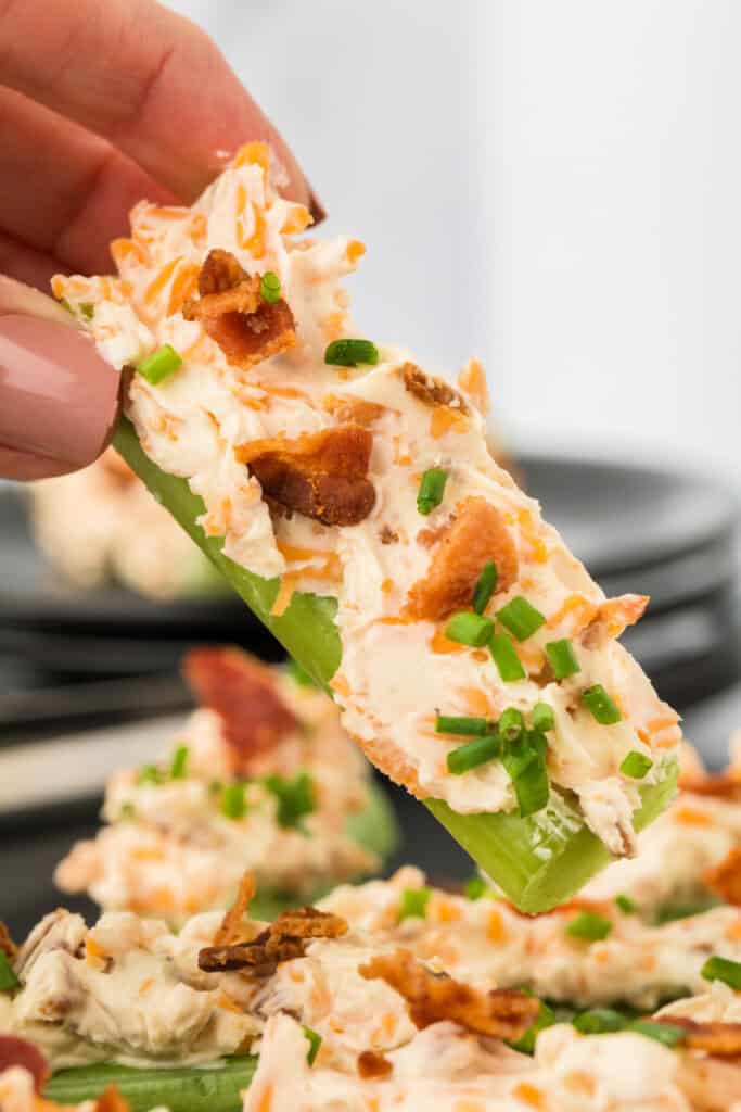 close-up of a hand holding a celery stick filled with cheese, bacon, and chives