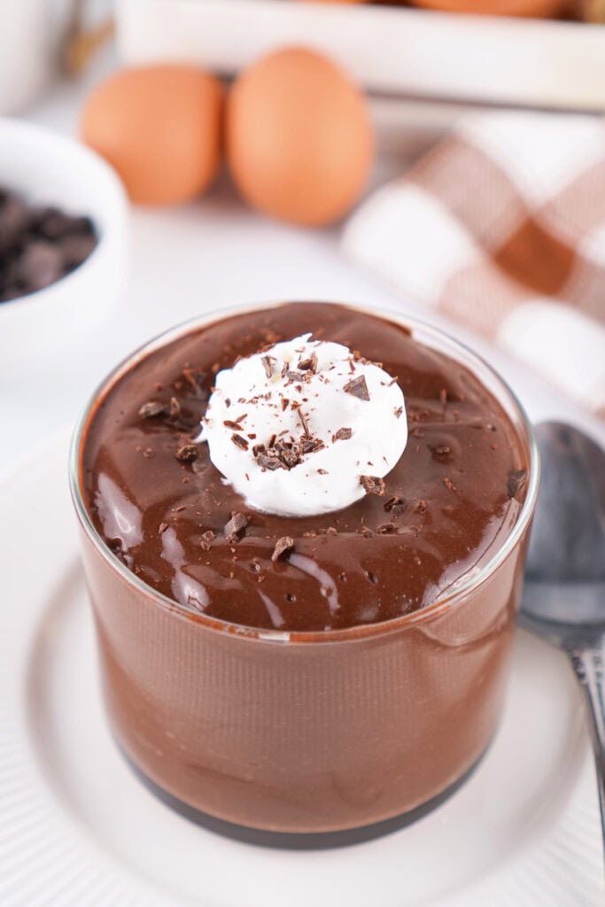 chocolate pudding in a glass topped with whipped topping and chocolate shavings with eggs in the background