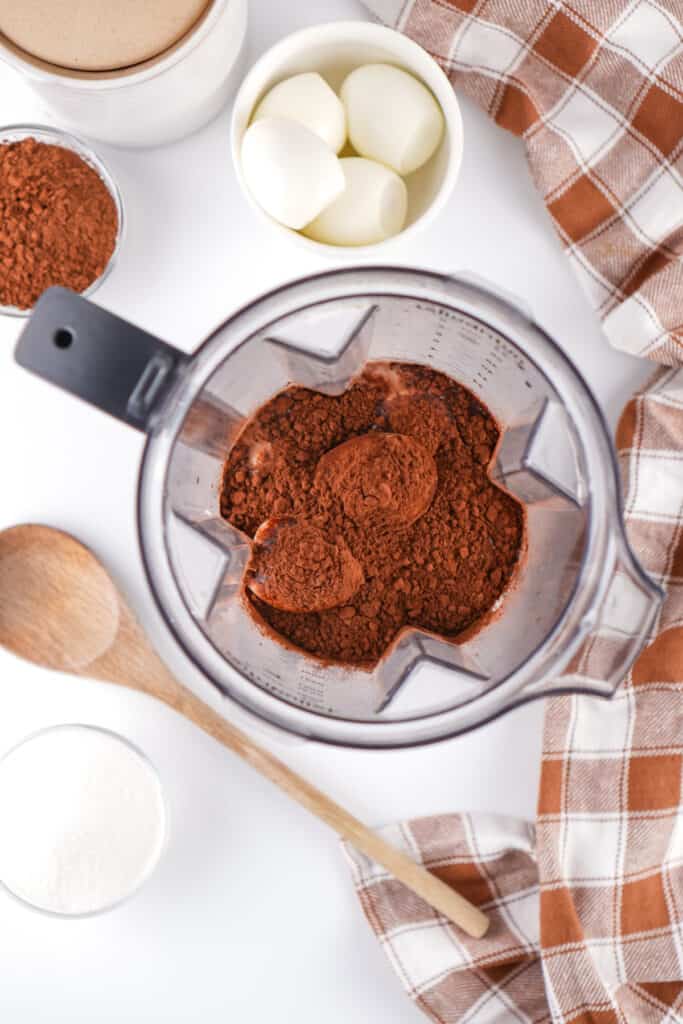 blender with cocoa powder and ingredients on a white surface with eggs and sugar nearby