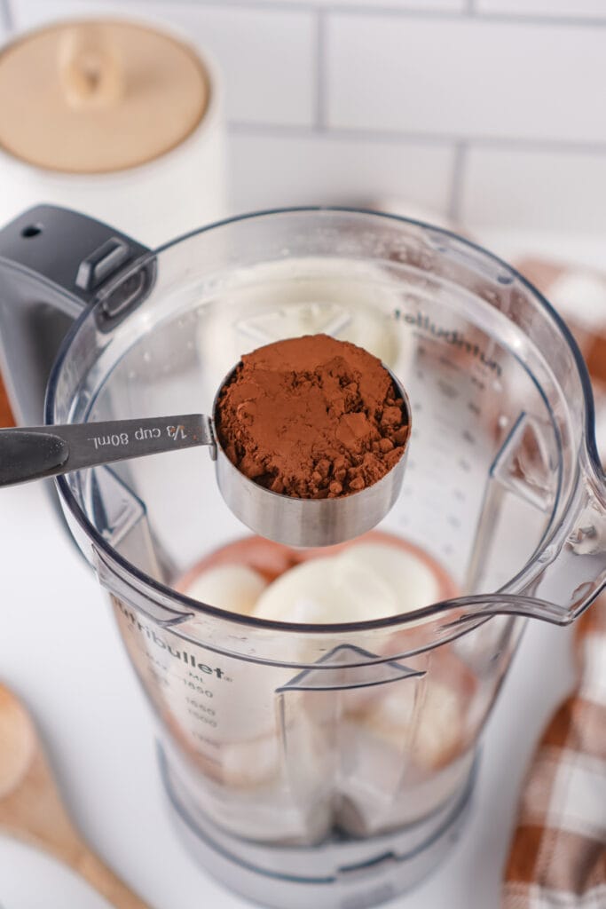 measuring cup of cocoa powder held over blender with hard boiled eggs inside