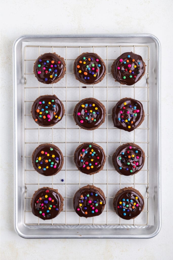 overhead view of chocolate cookies topped with ganache and rainbow candy-coated chips on a cooling rack