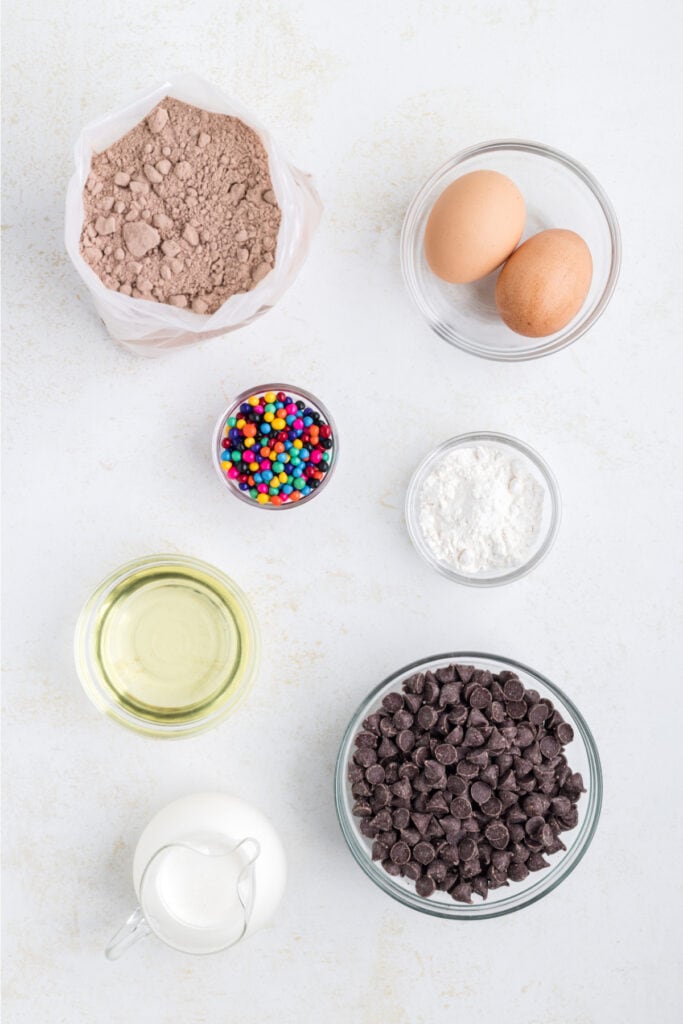 ingredients for chocolate cookies including brownie mix, eggs, oil, flour, chocolate chips, cream, and candy-coated chips