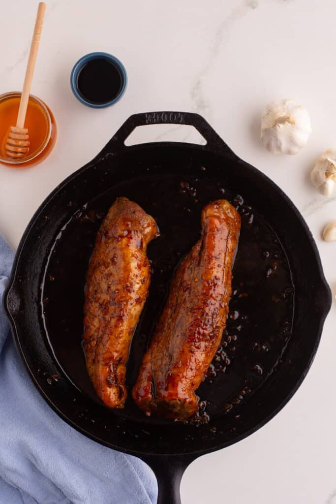 Honey garlic pork tenderloins coated in glaze in a cast iron skillet with honey jar and garlic cloves nearby.