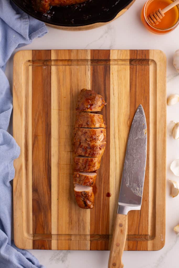 Sliced honey garlic pork tenderloin on a wooden cutting board with a chef knife beside it.