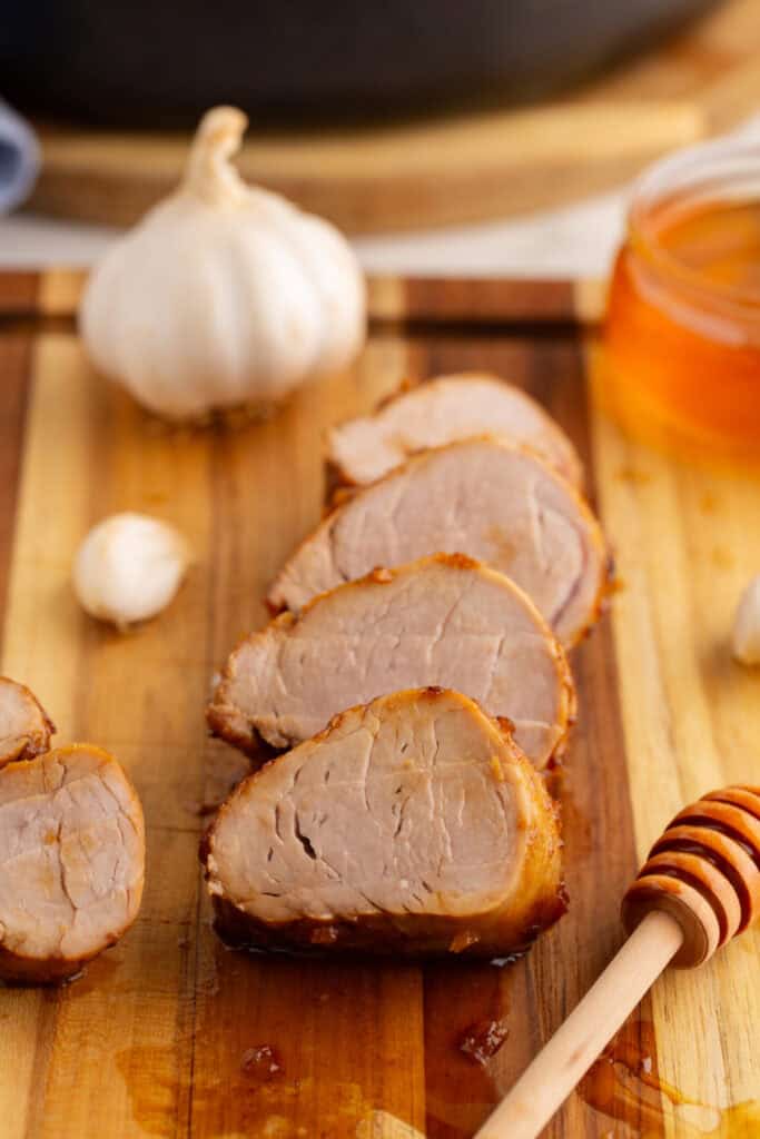 Sliced honey garlic pork tenderloin on a wooden cutting board with a garlic bulb and jar of honey in the background.