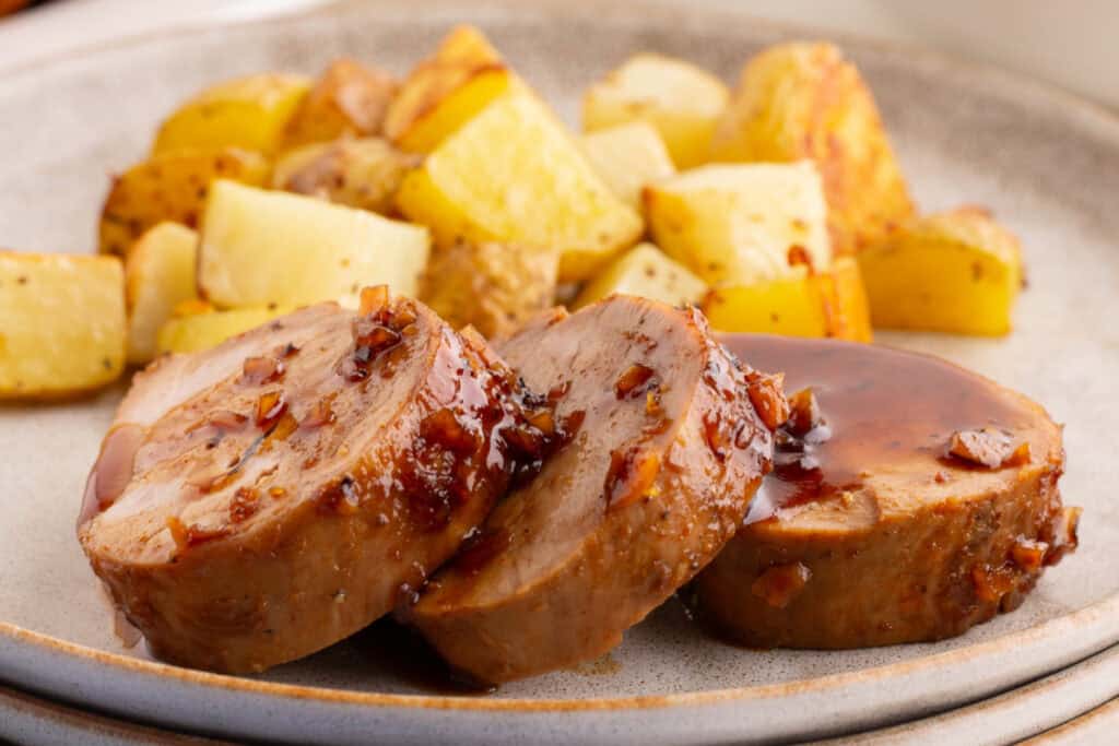 Honey garlic pork tenderloin slices on a plate with roasted potatoes in the background.