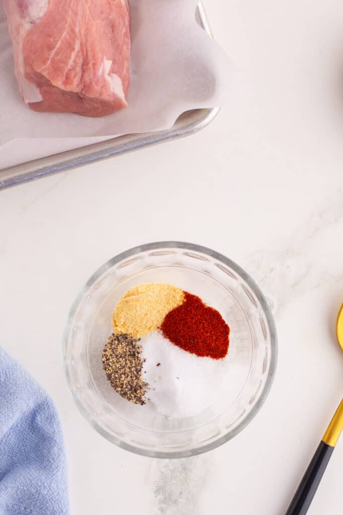 Raw pork tenderloin on parchment-lined baking sheet next to a small bowl with salt, pepper, paprika, and garlic powder.
