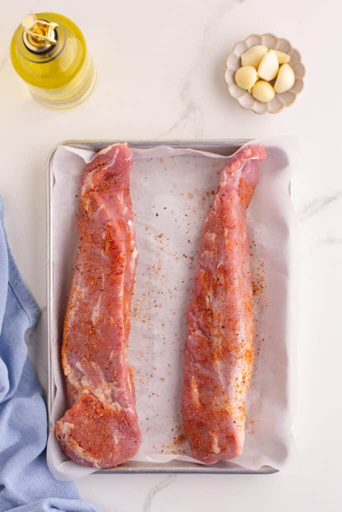 Two raw pork tenderloins on a parchment-lined baking sheet sprinkled with seasoning.