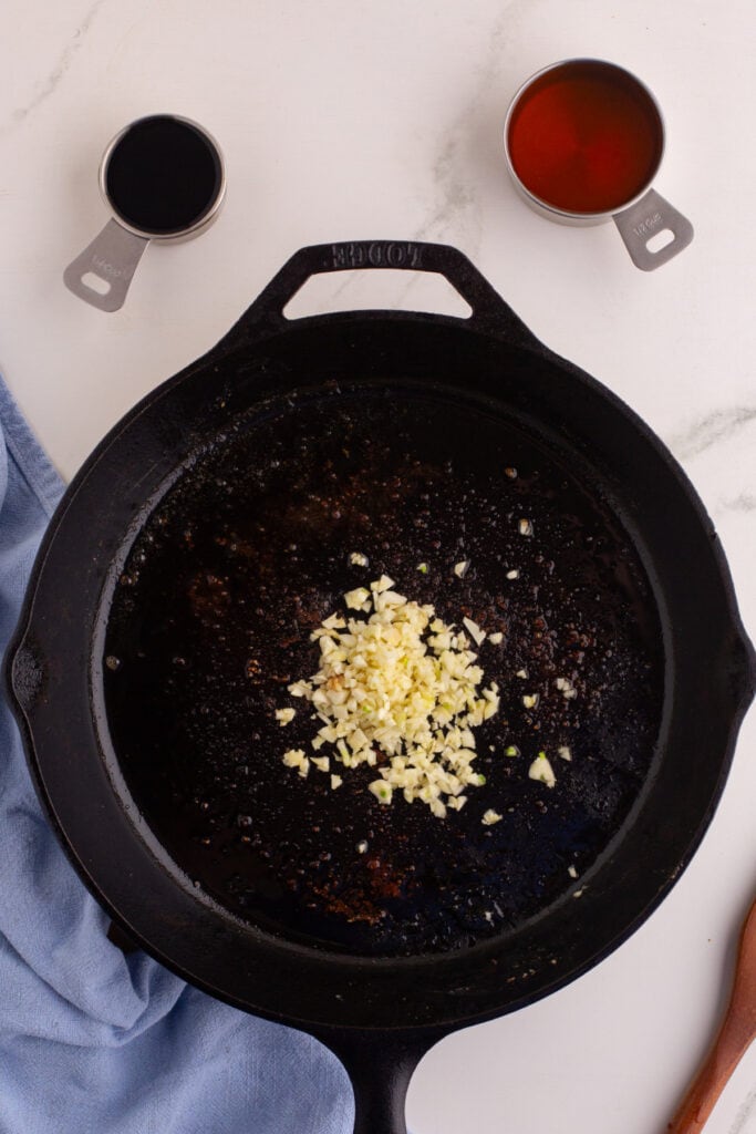 Chopped garlic cooking in a cast iron skillet with measuring cups of soy sauce and honey above the pan.