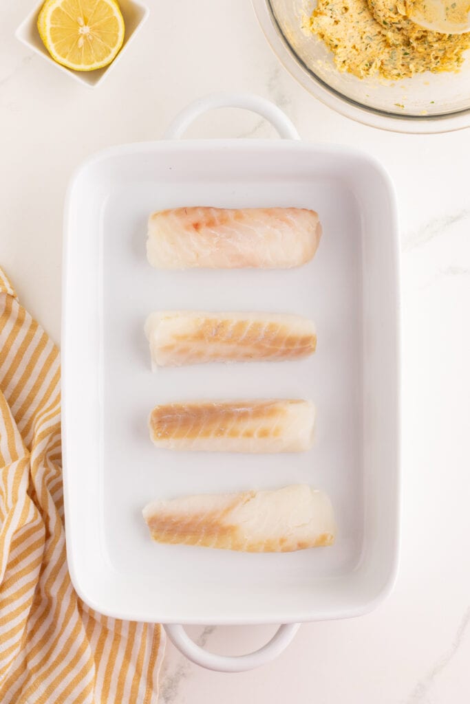 Raw cod fillets arranged in a white baking dish on a marble countertop.