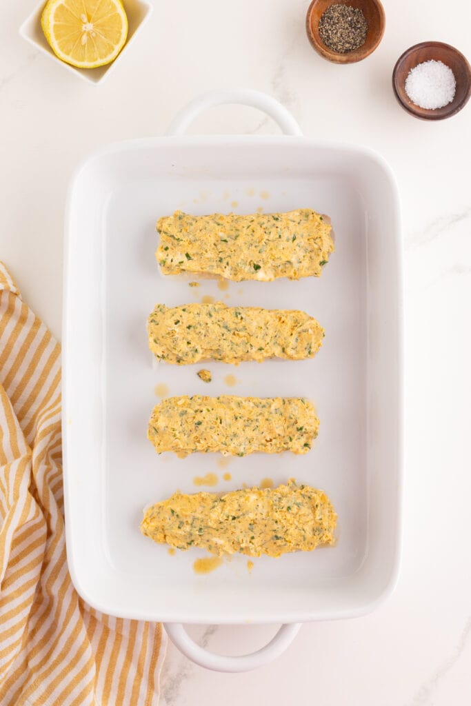 Cod fillets in a white baking dish spread with garlic herb butter mixture before baking.