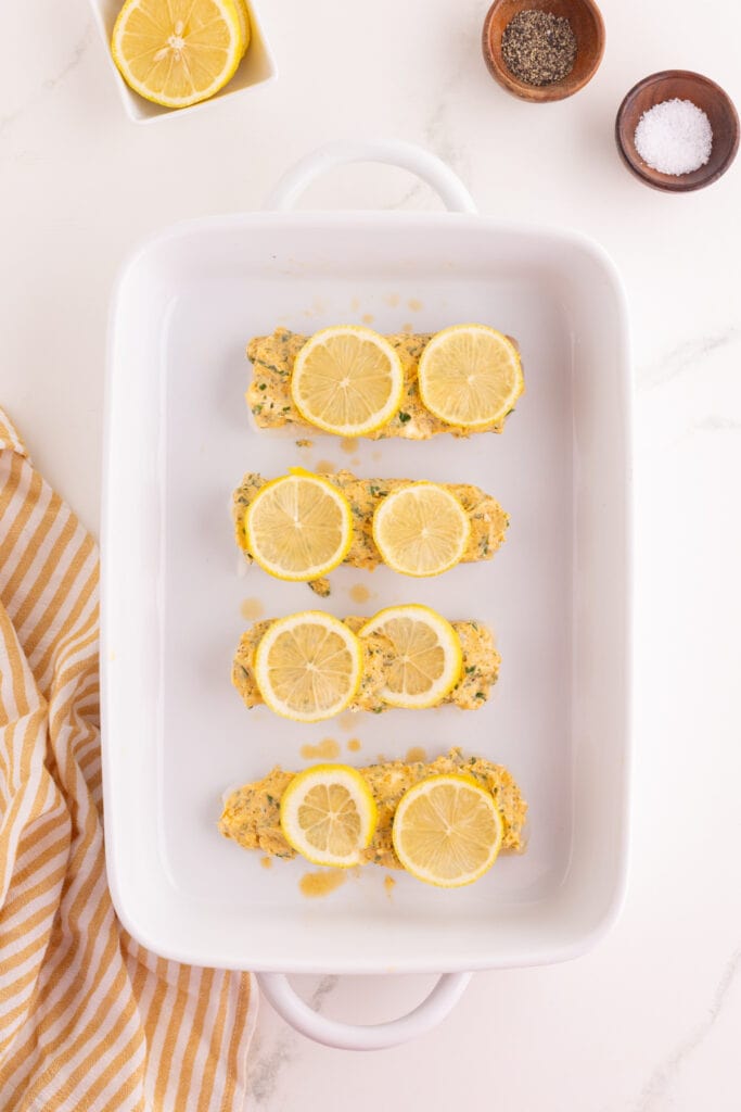 Cod fillets topped with lemon slices and garlic herb butter mixture in a white baking dish before baking.