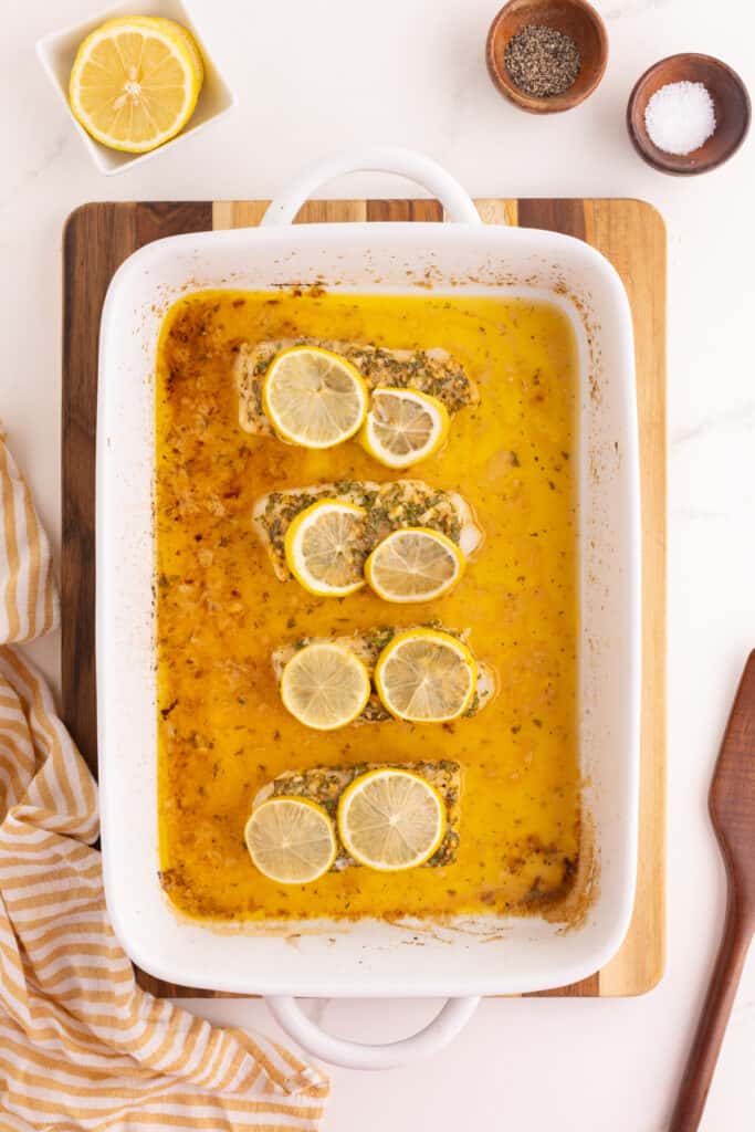 Baked cod fillets in a white baking dish surrounded by melted butter sauce with lemon slices on top.