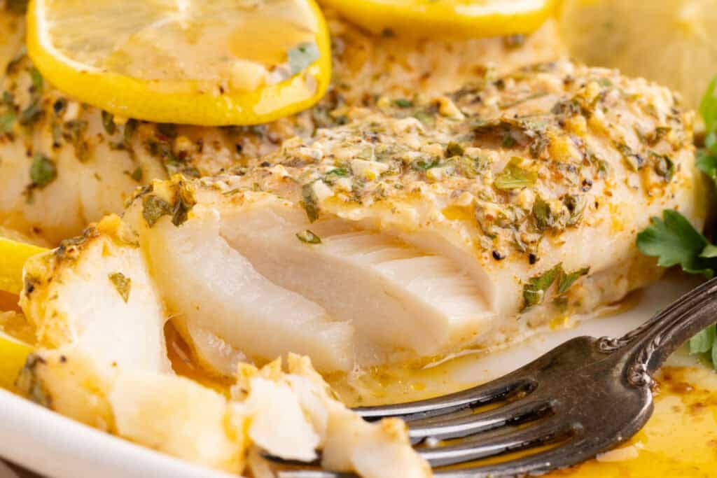 Close up of baked cod showing flaky white interior topped with garlic herb butter and lemon slices.