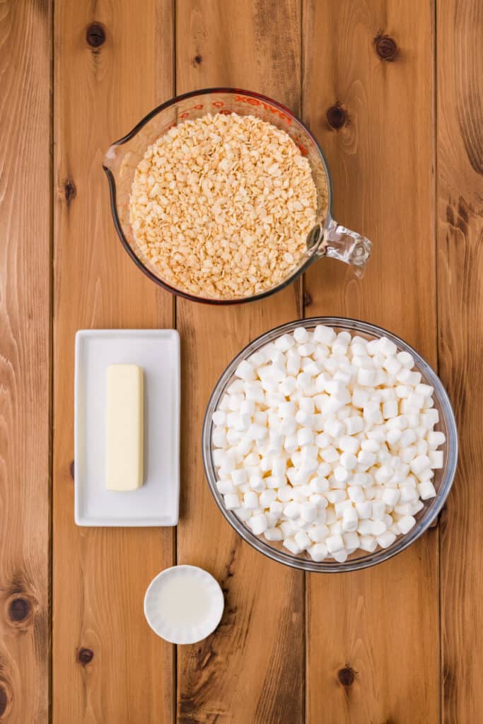 measuring cup filled with crispy rice cereal next to a bowl of mini marshmallows, stick of butter on a dish, and small bowl of lemon extract on a wooden surface