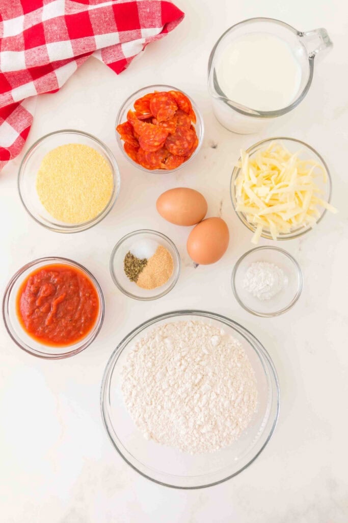 ingredients for pizza muffins including flour, cornmeal, pepperoni, shredded mozzarella, eggs, milk, pizza sauce, and seasonings arranged in bowls on a white surface