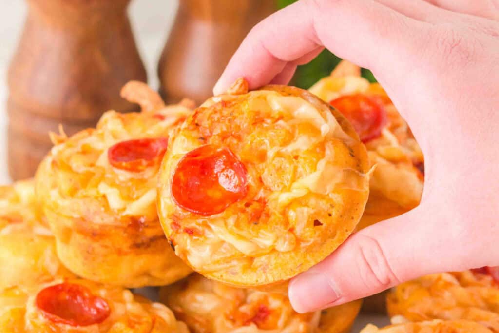 hand holding a baked pizza muffin with melted cheese and pepperoni over a tray of muffins