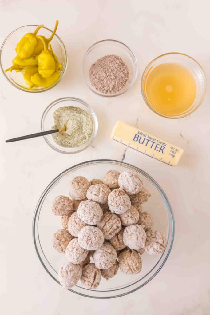 frozen meatballs in a glass bowl with pepperoncini, ranch seasoning, au jus gravy mix, chicken broth, and butter on a countertop