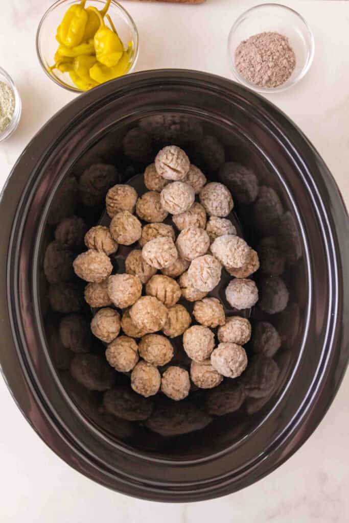 frozen meatballs placed inside a slow cooker with bowls of pepperoncini and seasoning nearby
