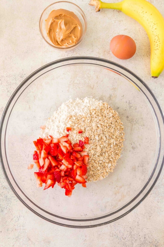 flour, oats, and chopped strawberries in a glass mixing bowl with banana, peanut butter, and egg nearby