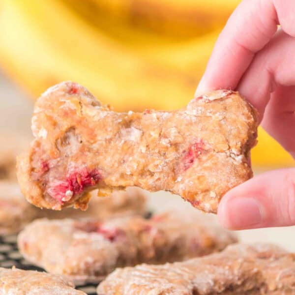 close-up of a hand holding a strawberry banana dog treat bone above baked dog treats on a cooling rack