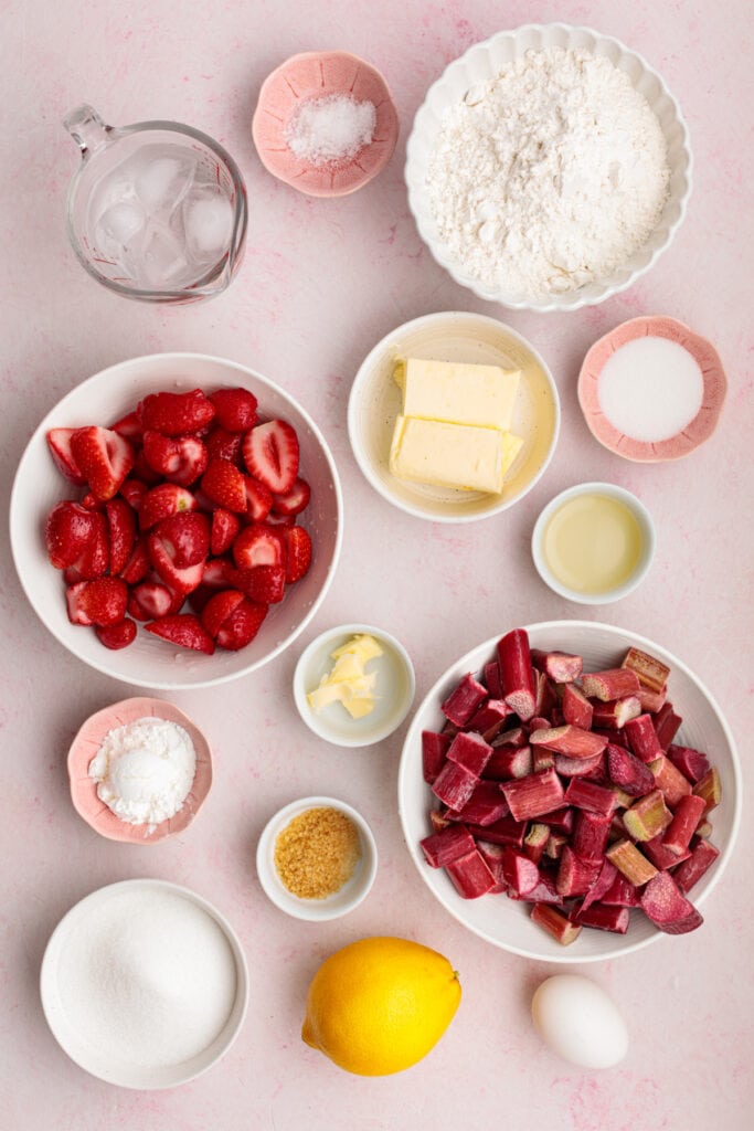overhead view of ingredients for strawberry rhubarb galette including sliced strawberries, chopped rhubarb, flour, butter, sugar, lemon, egg, and ice water on a pink background