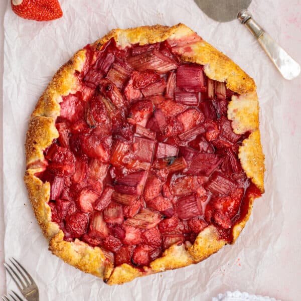 overhead view of baked strawberry rhubarb galette on parchment paper with a fork and pie server nearby