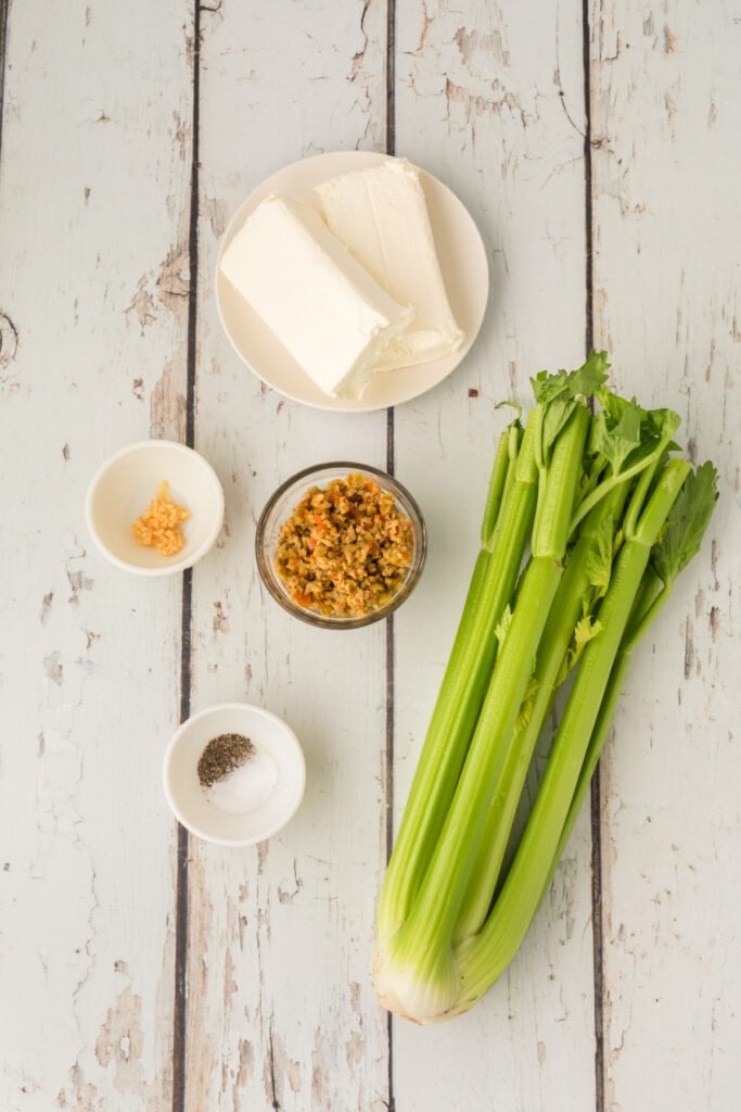 celery stalks, cream cheese blocks, chopped olives, minced garlic, and salt and pepper in small bowls on a wooden surface