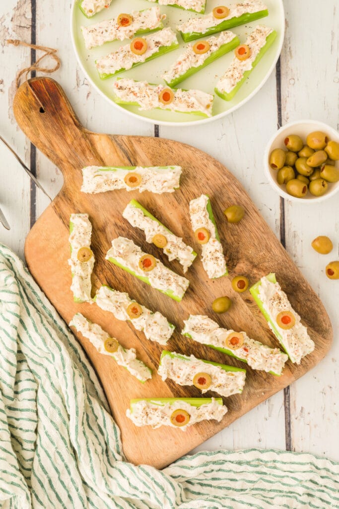 stuffed celery arranged on a wooden board with sliced olives and seasoning nearby
