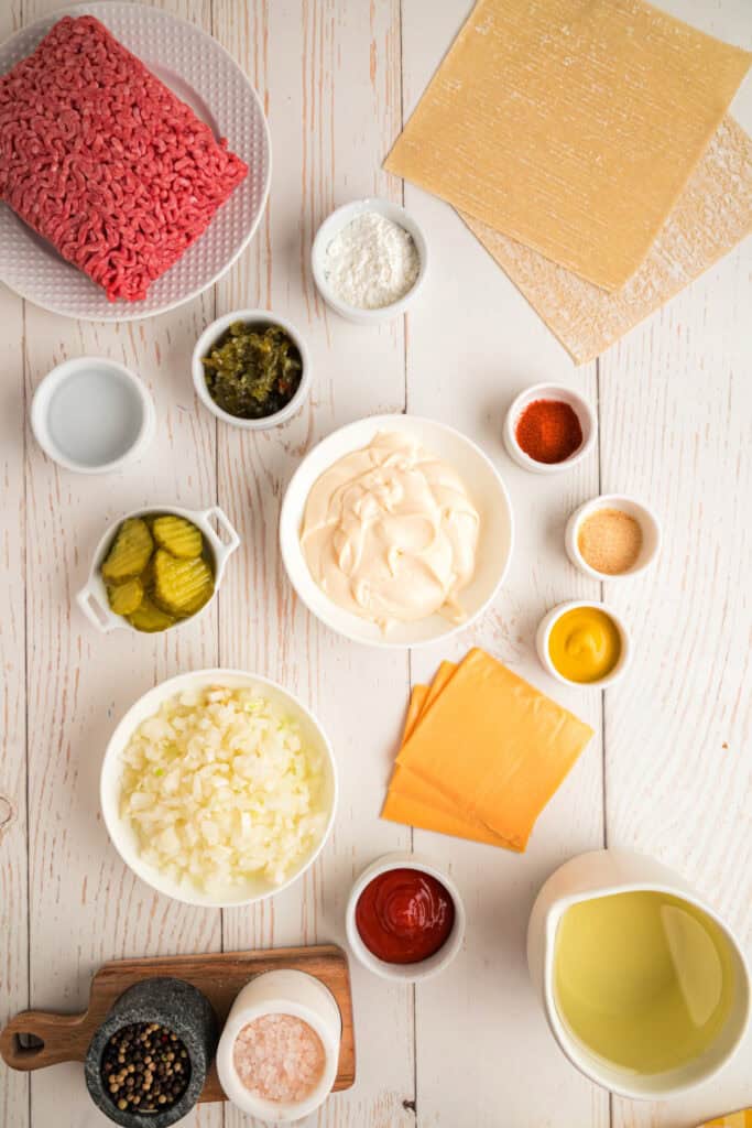 ingredients for Big Mac Egg Rolls arranged on a white wooden surface including ground beef, cheese, pickles, onion, and sauce