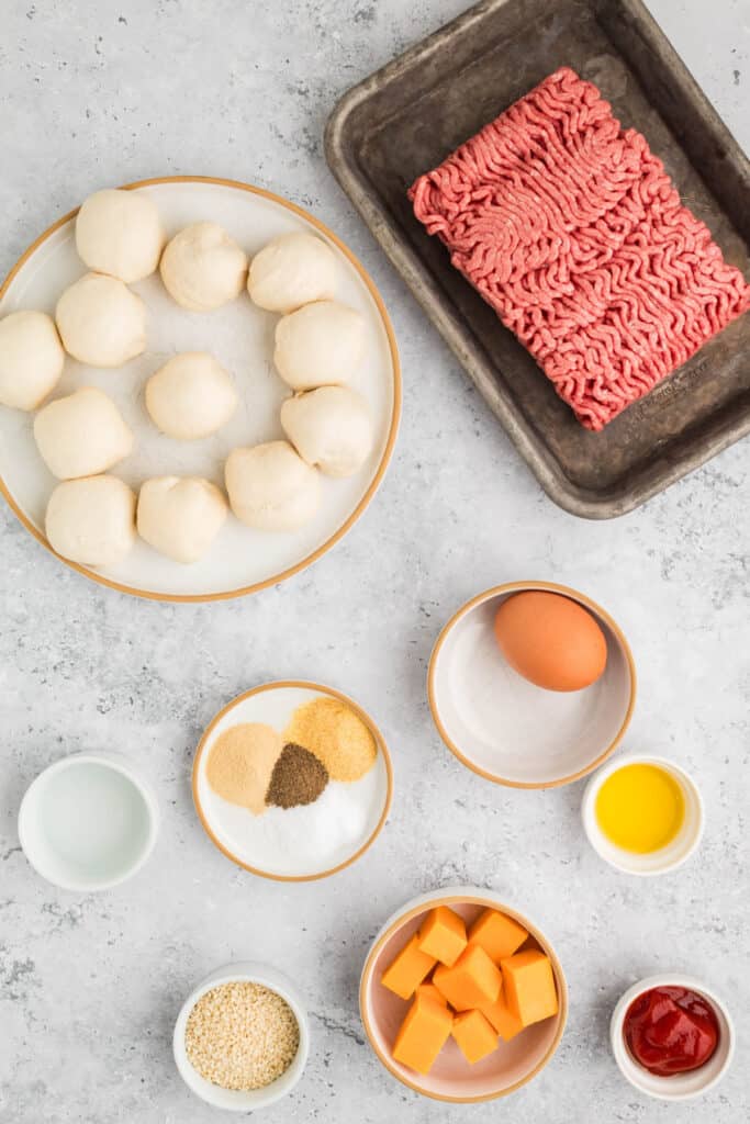raw ground beef on a tray with dough balls, cheese cubes, egg, ketchup, and seasonings arranged on a countertop