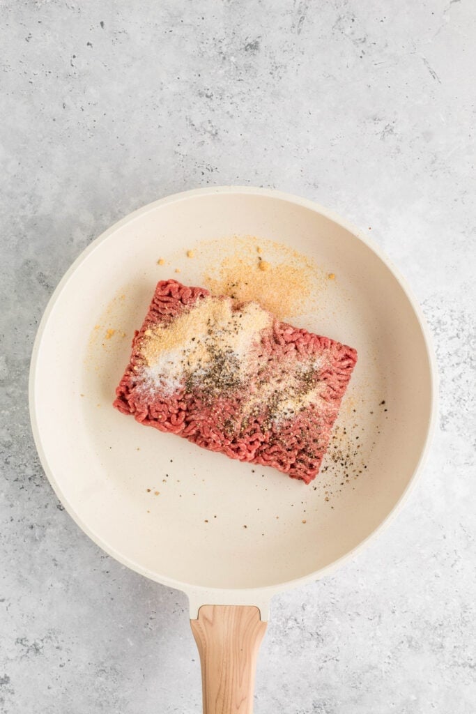 raw ground beef in a skillet with garlic powder, onion powder, salt, and pepper on top