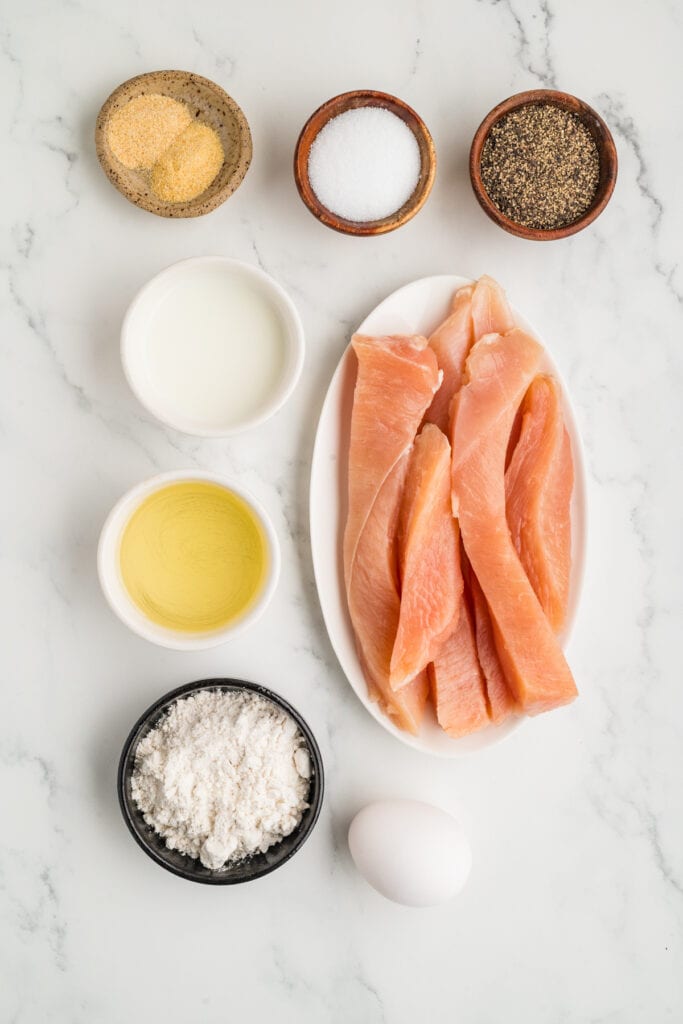 raw chicken strips on a plate with small bowls of flour, milk, oil, salt, pepper, and seasonings on a white surface