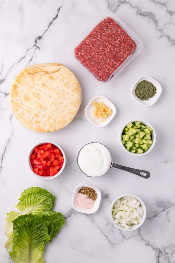 pita bread, raw ground beef, chopped tomatoes, cucumber, onion, garlic, yogurt, lettuce, and dried herbs on a marble surface