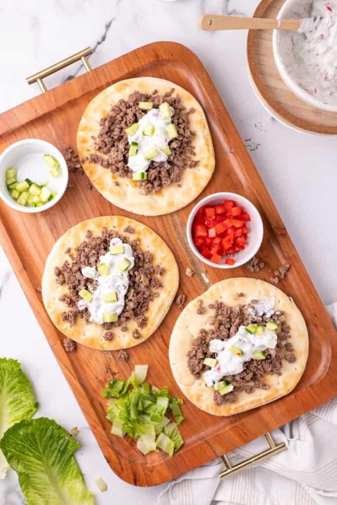 pita bread topped with ground beef, yogurt sauce, cucumber, tomato, and lettuce on a wooden tray