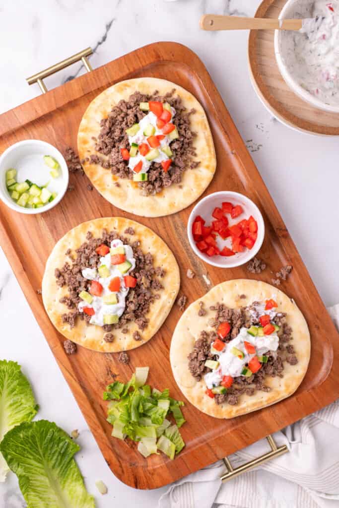pita breads topped with ground beef, yogurt sauce, and vegetables served with fries on a wooden tray