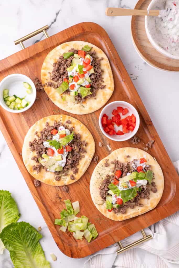close view of pita bread topped with ground beef, yogurt sauce, cucumber, tomato, and lettuce with fries on the side