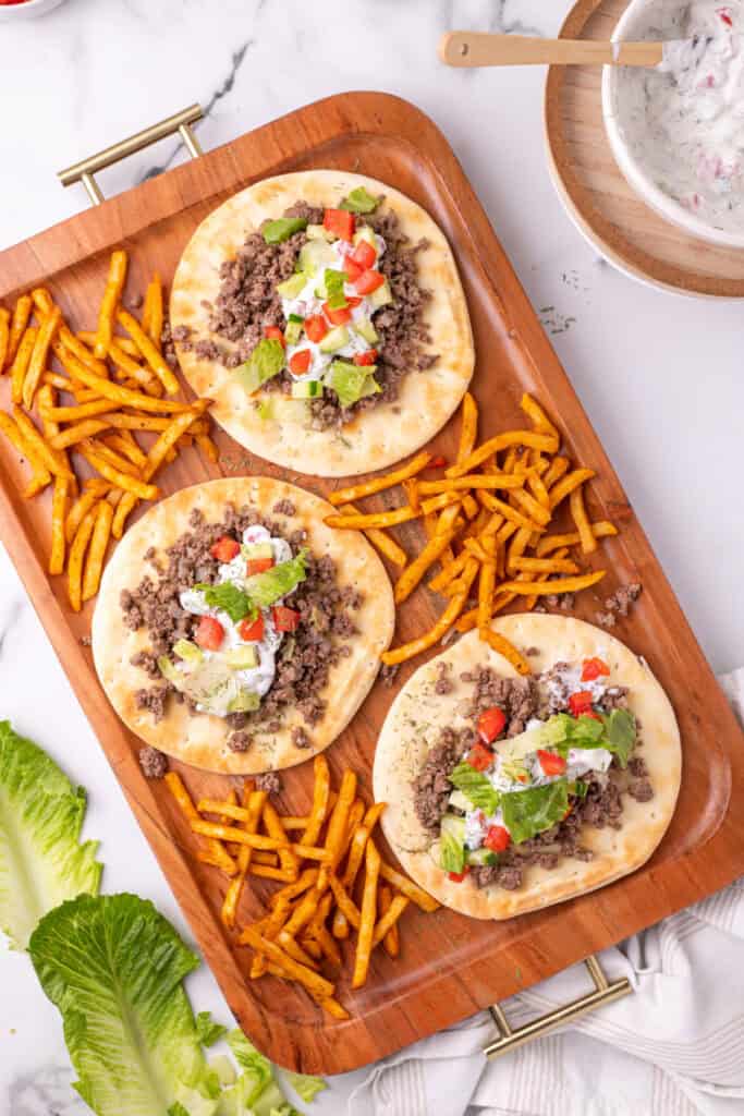 close view of pita bread topped with ground beef, yogurt sauce, cucumber, tomato, and lettuce on a wooden tray