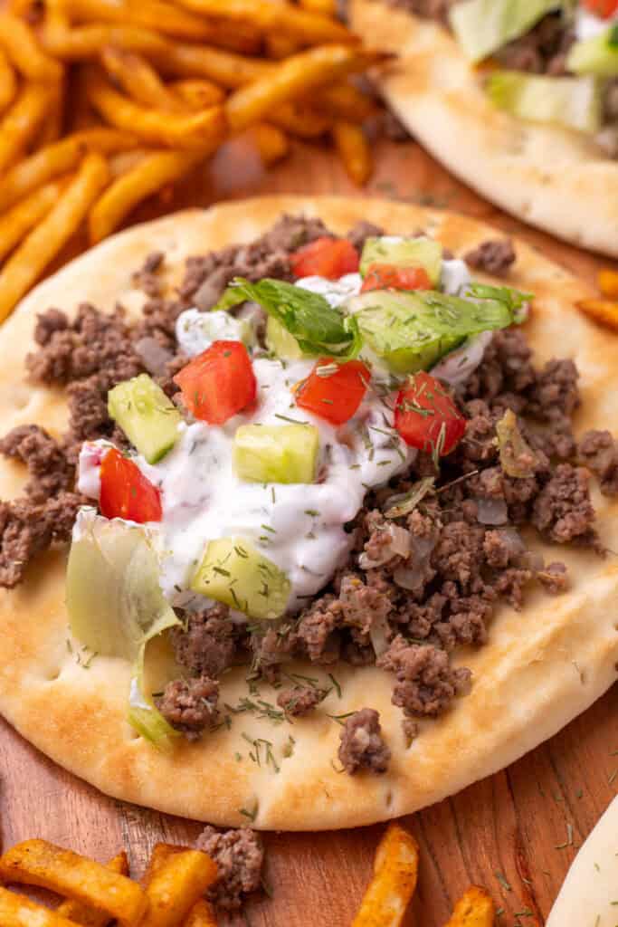 close view of pita bread topped with ground beef, yogurt sauce, cucumber, tomato, and lettuce with fries